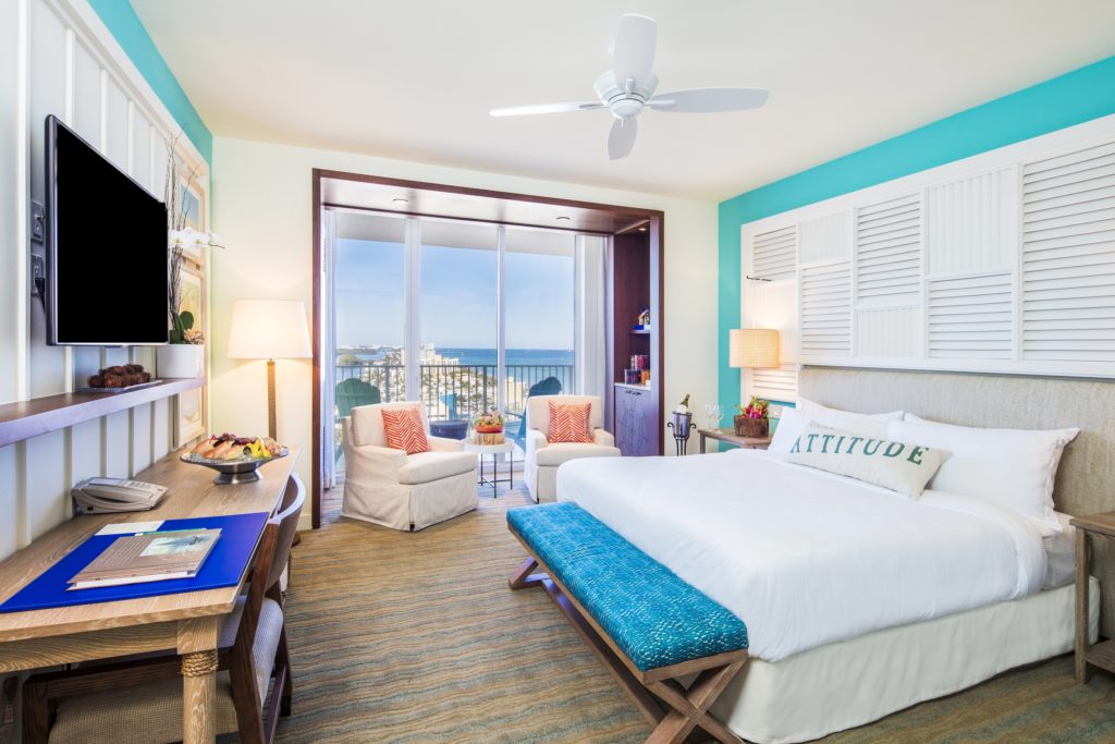 Room contemporary furnishings with a sea and sky color palette and king bed, two armchairs, table, tv, large window and balcony with partial views
