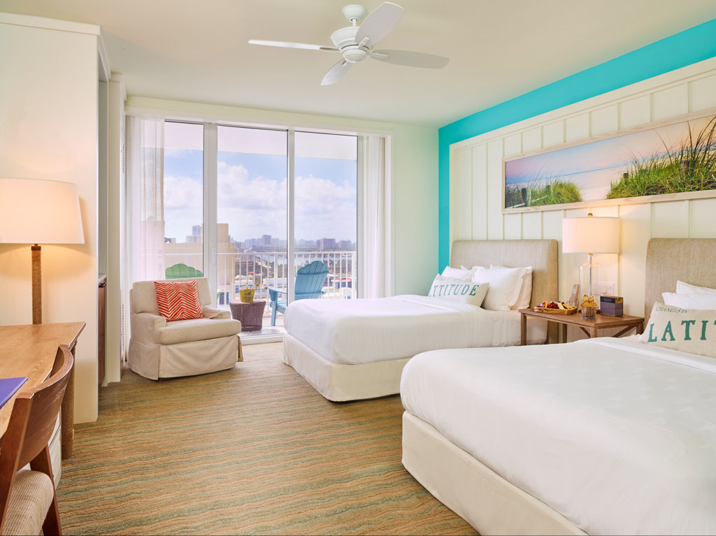 Room contemporary furnishings with a sea and sky color palette and two queen beds, armchair, table, large window and balcony with partial views