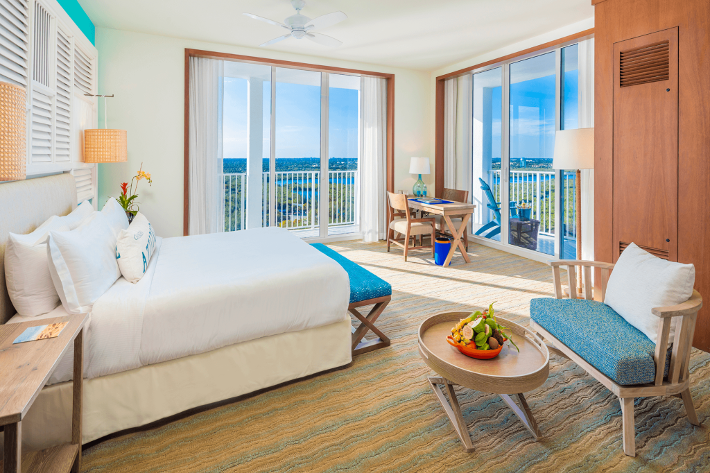 Room contemporary furnishings with a sea and sky color palette, one king bed, armchair, table and two large windows and balcony