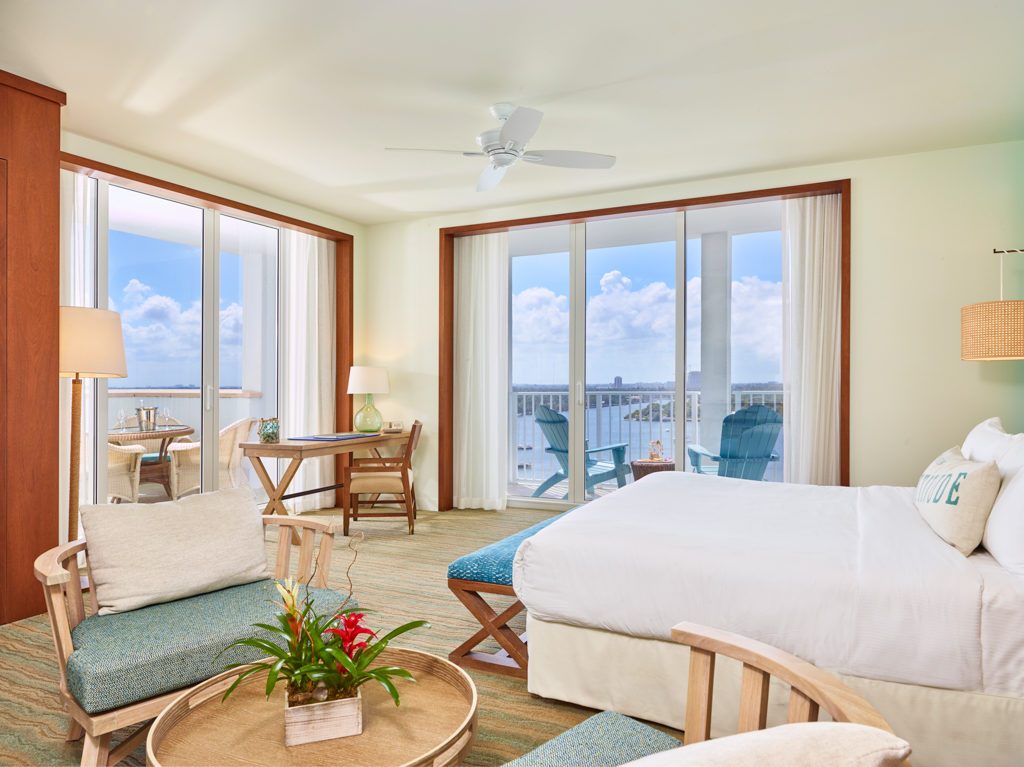 Room contemporary furnishings with a sea and sky color palette, one king bed, armchair, table and two large windows and veranda